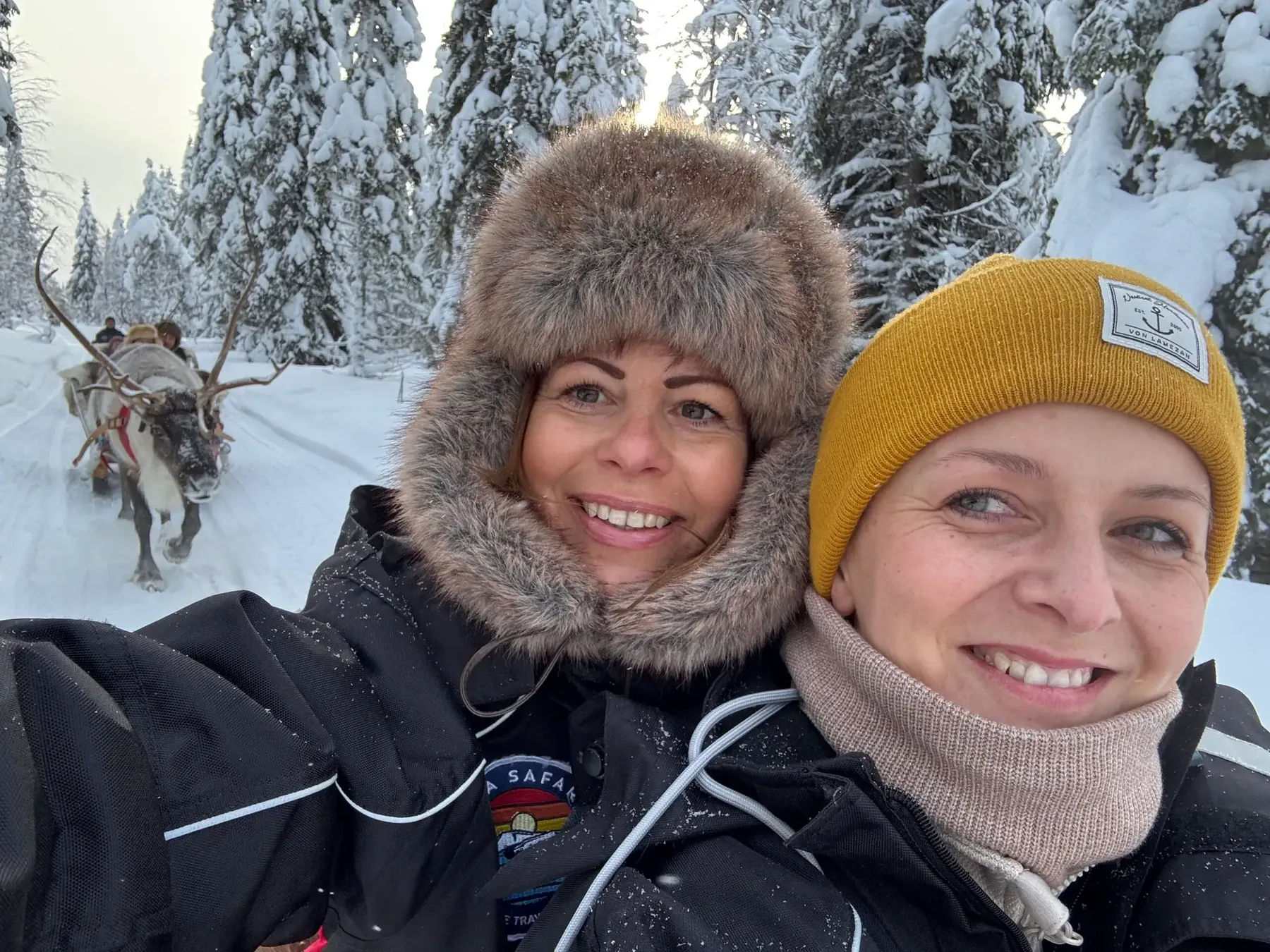 Zwei Reisebegleiterinnen im Schnee, hinter ihnen ein Rentiergespann